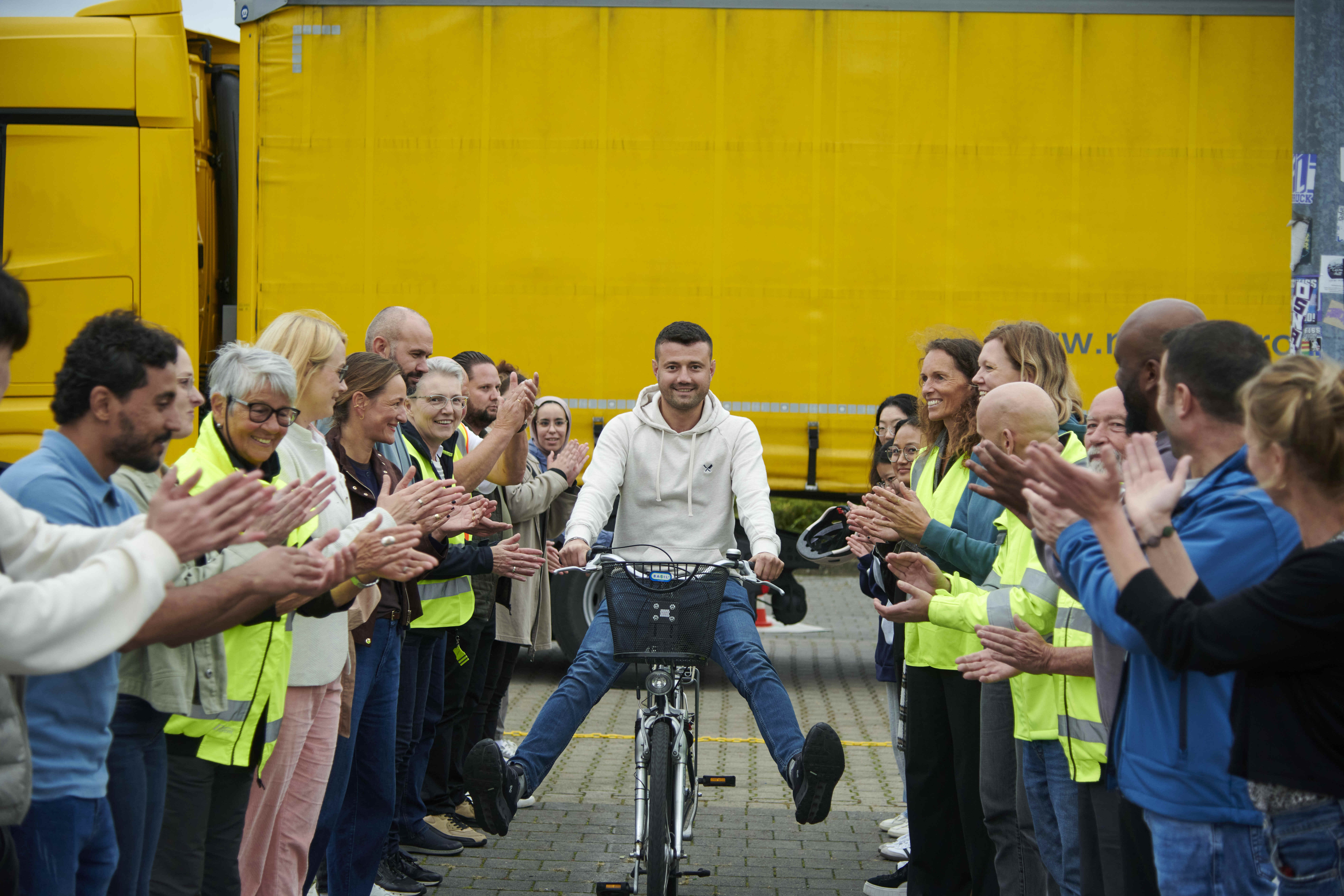 Ein Mann sitzt lächelnd auf einem Fahrrad und wird von einer Gruppe applaudierender Menschen in Warnwesten empfangen. Im Hintergrund steht ein großer gelber Lkw.